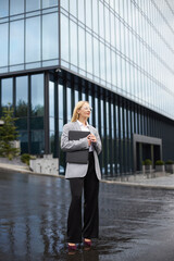 Pensive middle aged blonde businesswoman holding laptop, looking away, waiting someone on the street, rainy day