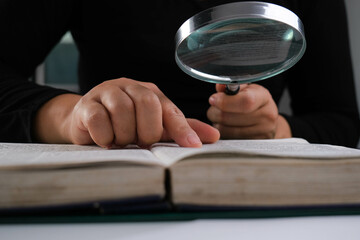 Close-up of a woman looking through a magnifying glass at a textbook. Magnifying glass in hand and open book on table. Education and research concept. - Powered by Adobe