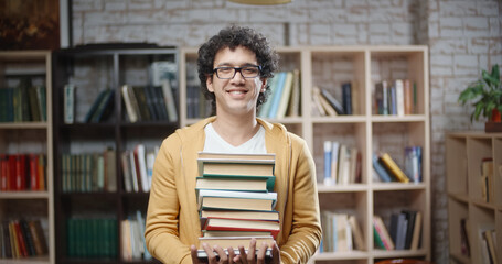 Asian guy with curly hair is standing between bookshelves of library holding books in hands, looking at camera and smiling - portrait closeup, education, student lifestyle concept 