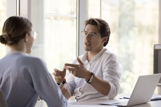 Serious handsome young business professional man talking to female colleague at workplace table. Mentor teaching student, intern, new employee. Company boss interviewing job candidate