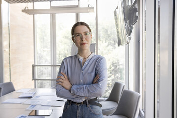 Positive confident young businesswoman in glasses posing in office interior, standing at meeting table with arms folded, looking at camera. Female business leader professional portrait