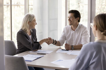 Happy mature business leader woman shaking hands with colleague, partner over paper marketing report. Female boss expressing recognition to employee, giving handshake, smiling
