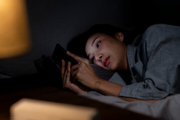 Young Asian woman using smartphone in bed cannot sleep from insomnia, depressed woman in bedroom