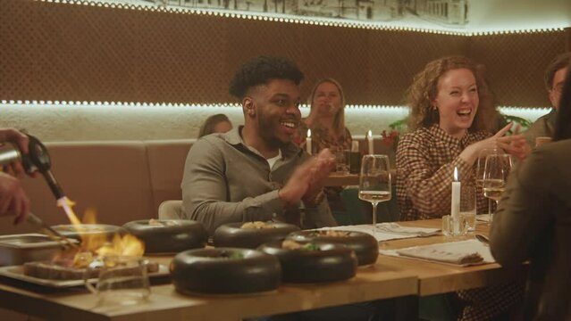 The chef prepares meal in flambe style in front of people in modern gastro cafe. Cheerful multi ethnic friends spend weekend evening, hangout on birthday party in restaurant. Concept of public eating.