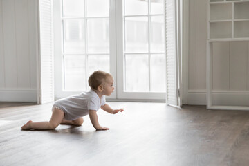 Adorable baby wearing white bodysuit, crawling on knees on floor at home. Curious active little infant child learning to move on warm heating safe floor, passing by window in background - obrazy, fototapety, plakaty