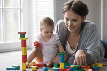 Happy young new mom teaching few month baby to build tower from colorful toy pieces, playing with infant child in diaper sitting on heating floor, having fun, training kid skills