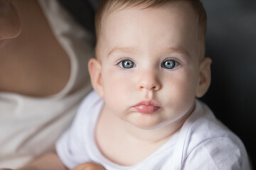 Face of pretty adorable blue eyed baby held by mom looking at camera. New mother holding little kid with tenderness, love, care, enjoying childcare. Beautiful infant indoor closeup portrait