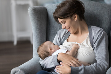 Happy young mother feeding baby mild from breast, looking at adorable sleepy kid with love,...