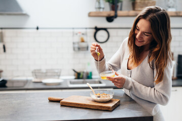 Young woman in the kitchen adds honey to muesli. - 590709978