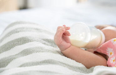 Hand of newborn baby holding and drinking milk from bottle alone on bed at home