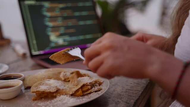 Young woman developer has lunch break eating food while working on laptop in outdoor cafe. Female freelancer having pankakes and coffee at coffeeshop for breakfast. IT girl eats her food at cafe