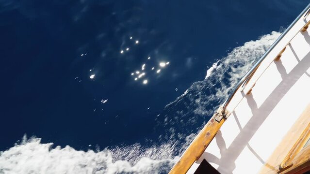 A view from above of the deck and side of a wooden boat sailing on the sea. The ship splits the brilliant foamy white waves of the deep blue sea. Slow motion