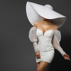 Fashion Woman in Big White Hat hiding Face. Elegant Bride in Mini Dress. Beautiful Lady in Wedding Gown. Stylish Glamour Girl in Sexy Party Dress over Gray Background - 590719523