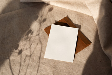 Blank paper card, envelope on neutral beige background, harsh sunlight floral shadows on sunset....