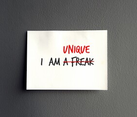 Note paper on gray wall background with handwritten text I AM A FREAK, crossed off to I AM UNIQUE - concept of self respect, to overcome negative self talk by accept yourself with love and care