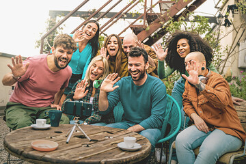 Diverse Friends in a Video Call Together: Seven friends sitting around a wooden table, smiling and waving at a smartphone on a tripod, having a video call.