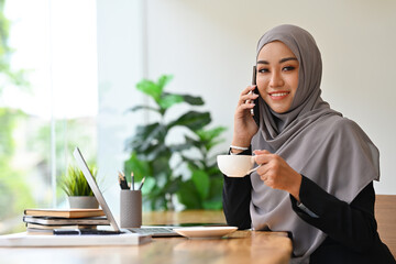 Pleasant young muslim woman entrepreneur drinking coffee and talking on mobile phone.