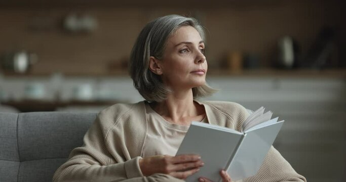 Peaceful positive petty mature woman reading book at home, turning head away, thinking, smiling, enjoying bestseller novel, leisure time, studying new knowledge, skills, feeling relaxed