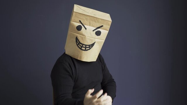 A man in a black turtleneck with a bag on his head, with an angry smiley face drawn, emotionally gloats while sitting on a chair. Emotions and gestures.