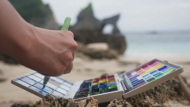Plein-air lifestyle. Close-up of watercolor palette and hand of artist mixing paints while painting. Drawing outdoors on a beautiful beach in the wild nature. Watercolor painter and her tools. Summer.