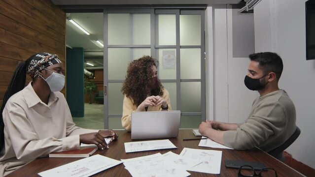 Full shot of three multiethnic colleagues sitting in conference room in modern coworking office, with protective masks on faces, and having animated discussion
