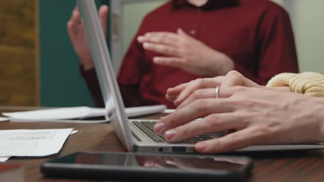 Close-up shot of hands of unrecognizable fair-skinned woman swiftly typing on laptop keyboard during business meeting in conference room, and anonymous colleagues talking in background