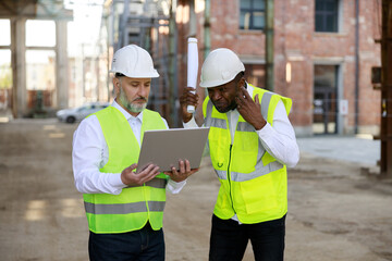 Caucasian senior foreman with wireless laptop discussing about all peculiarities of project to african american male architect at construction area. Concept of business meeting and building process.