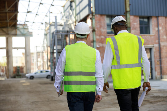 Back view of builder and architect inspecting building area while walking at construction site. Multiethnic partners wearing hardhats and reflective vest for protecting from accidents.