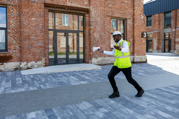 Happy african american builder with megaphone in hand dancing at construction site. Joyful foreman in vest and hardhat enjoying with successful work. Achievement and people concept.