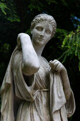 Vertical shot of a white marble statue of a young woman against the green trees