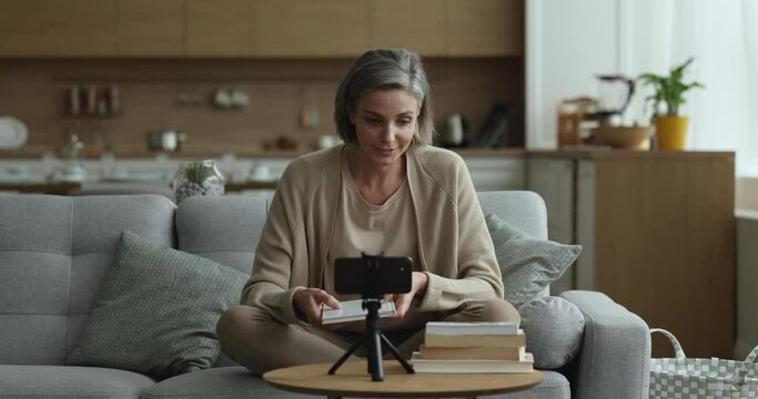 Pretty middle aged literature blogger presenting stack of bestsellers to blog audience, waving greeting hand hello at smartphone camera, speaking, showing book, fiving review, feedback