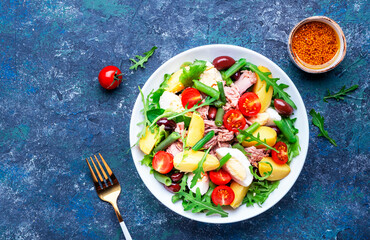 Nicoise salad with tuna, tomatoes, eggs, green beans, potatoes and olives on plate, blue table background, top view