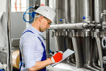 A young worker in a chemical factory makes notes in a notebook. Stainless steel metal structures...