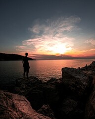 Image of a boy standing on the rocks looking at the sea and dark red sunset.