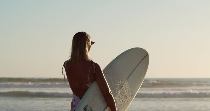 Slow-motion of a female surfer with a white surfboard in her hand looking at the waves of the ocean