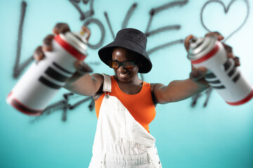 African Hipster girl painting graffiti on teal wall holding spray bottles wearing orange sunglasses