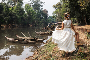 Woman wearing white robe dress posing near river with old wooden boats