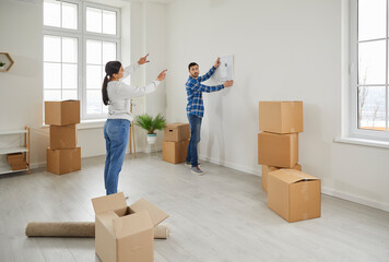 Young couple homeowners moving to new home and hanging photo or picture on the wall in their house. Man and woman decorating their new apartment. Relocating, real estate or mortgage concept.