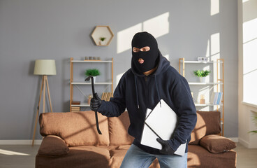 Thief with black balaclava making burglary, stealing two modern expensive laptops and running away....