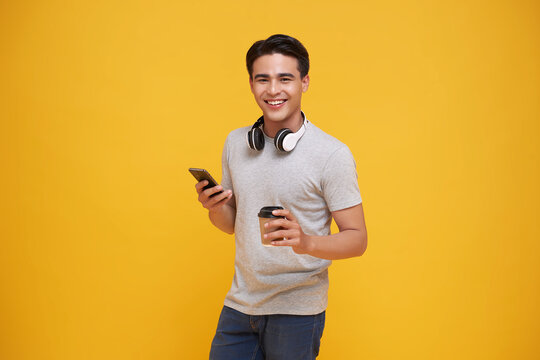 Happy Asian teen man wearing headphones listening to music and holding mobile phone with coffee on yellow studio background. People lifestyle relax