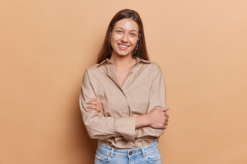 Studio shot of brunette young European woman smiles gently keeps arms folded wears brown shirt and...