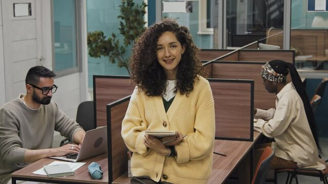 Medium full portrait shot of young Caucasian female freelancer posing in shared workspace facility, holding notepad and smiling, and diverse other customers sitting in cubicles and working on laptops