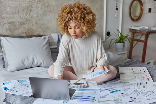 Busy woman focused at laptop screen does financial paper work at home checks bills plans budget studies accounting expenses dressed in domestic clothes sits on bed in bedroom cheks information
