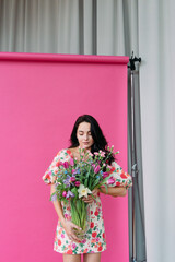 Young woman poses in studio with flowers in vase against pink photographic background.