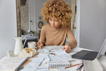 Curly haired woman sits at desk at home manages household budget studies financial bills and expenditures wears casual brown jumper plans expenses poses against domestic interior. Paper work concept