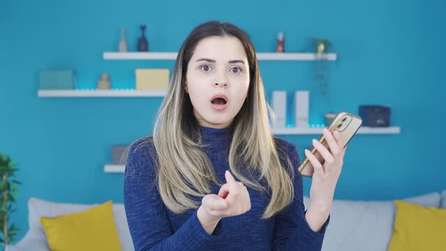 Young woman surprised and amazed by what she sees on the phone.
The young woman looking at her smartphone at home is confused and can't believe what she sees on the screen.
