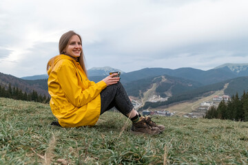 Young woman sits on hillside, drinking tea and smiling. Portrait of tourist girl in yellow jacket on the mountains
