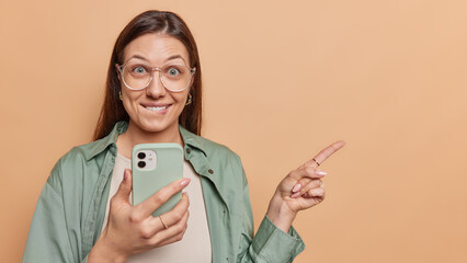 Cheerful surprised brunette young woman bites lips has wondered expression holds mobile phone points finger right on blank space wears green shirt isolated over brown background. Hey look there - obrazy, fototapety, plakaty