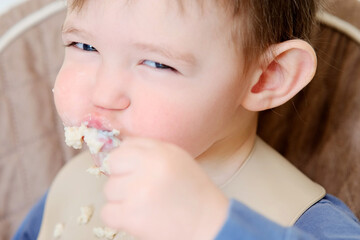A happy child eats porridge with a spoon while sitting on a high chair. Baby in a bib eats oatmeal porridge himself. Kid aged about two years (age one year nine months)