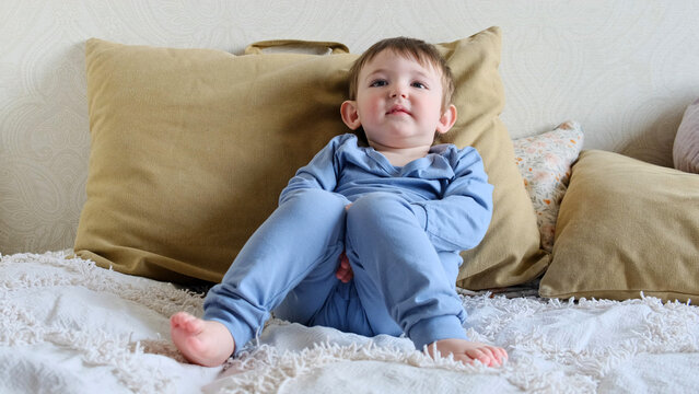 Baby in a blue clothes iaper sits on a sofa and watches TV. Child is resting sitting in a pajamas on a bed with pillows. Kid about two years old (age one year nine months)